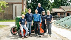 KI generiert: Eine Gruppe von sechs Personen posiert lächelnd bei sonnigem Wetter vor einem landwirtschaftlichen Gebäude neben einem grünen Traktor. Sie tragen überwiegend dunkelblaue Kleidung und scheinen Teil eines Teams oder einer Gemeinschaft zu sein.